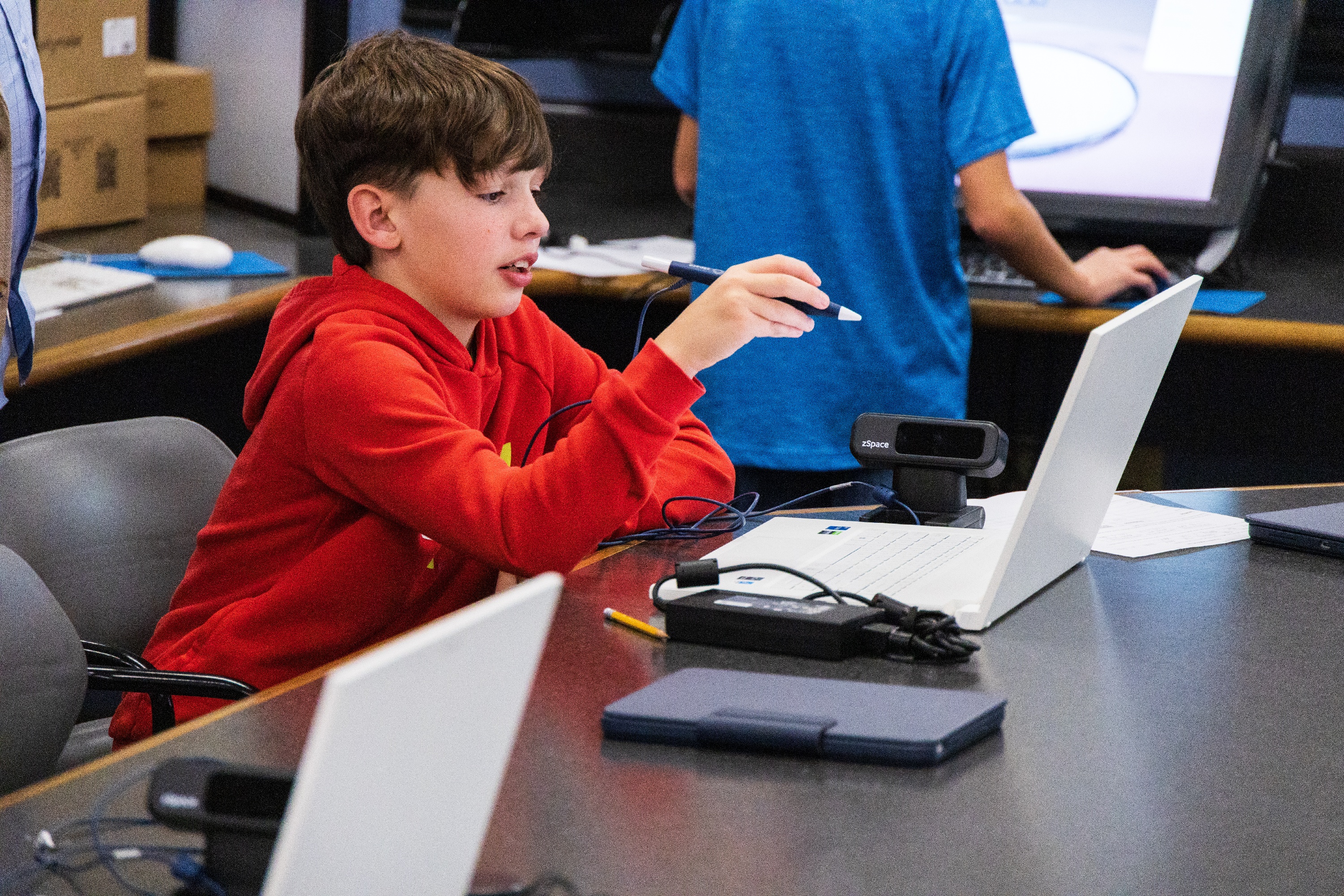 Middle school student in a red hoodie using a stylus pen with a zSpace virtual reality system in a classroom, highlighting interactive and immersive learning technology.