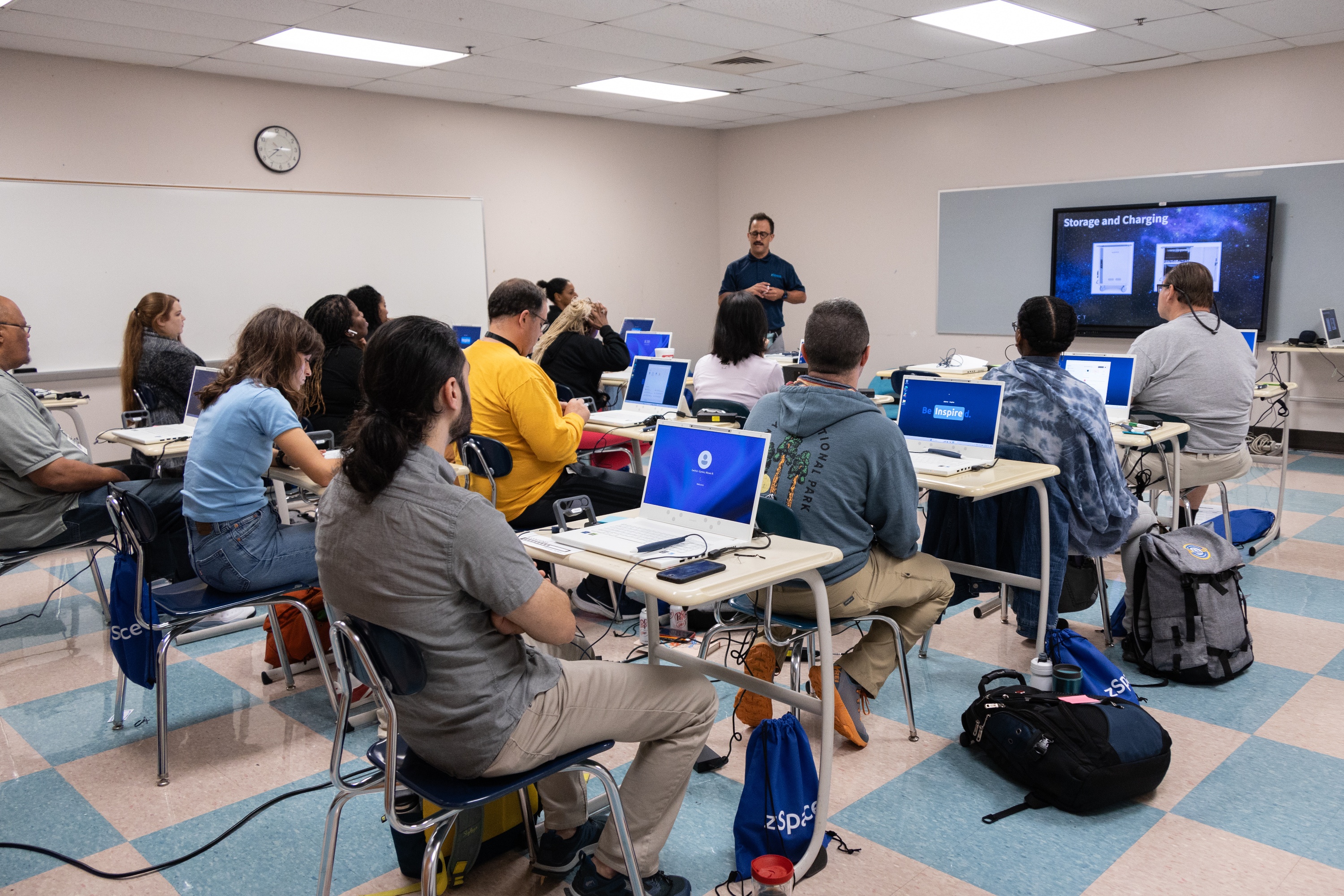 School teachers in a classroom seated at desks, each using a laptop, while the instructor stands at the front presenting 'Storage and Charging' information on a large screen.