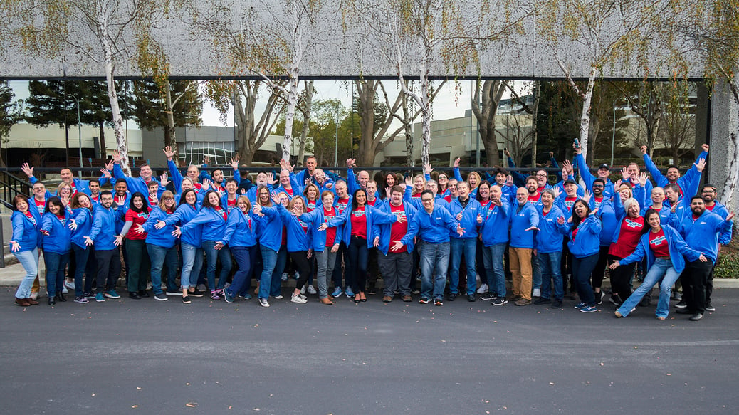 Large group of zSpace employees wearing blue jackets and red shirts standing outside in front of a building with large windows and trees. They are raising their arms in celebration, capturing a moment of unity and enthusiasm.