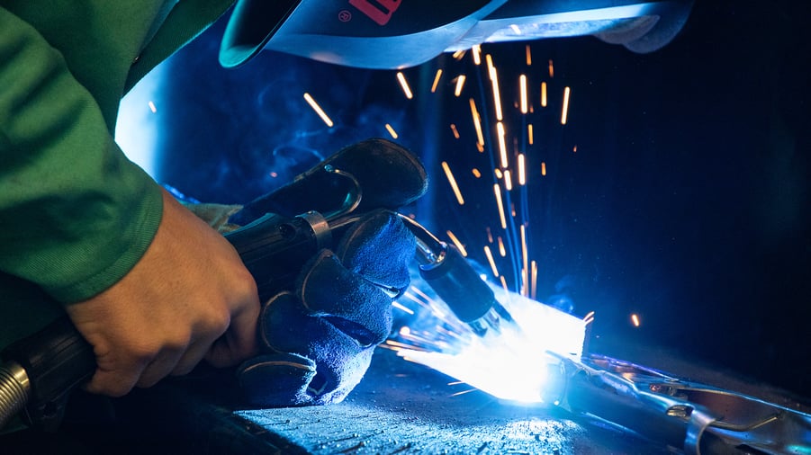 Close-up of a welder using a welding torch, with bright blue light and sparks flying. The welder is wearing protective gloves and a helmet, emphasizing safety in welding practices. This image highlights the precision and skill involved in welding, relevant for discussions on technical education and vocational training.