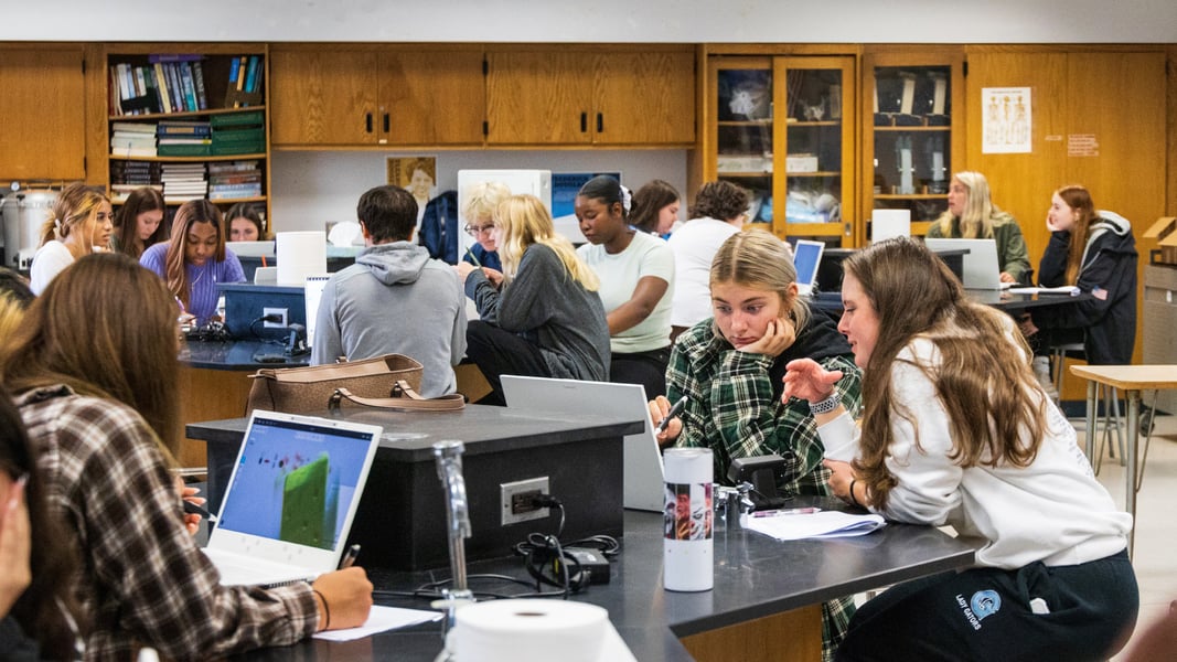 Students engaged in a collaborative learning environment in a science classroom, using zSpace laptops and lab equipment to conduct experiments and research. The classroom features wooden cabinets filled with books and scientific materials, highlighting the use of technology and hands-on learning in modern education.