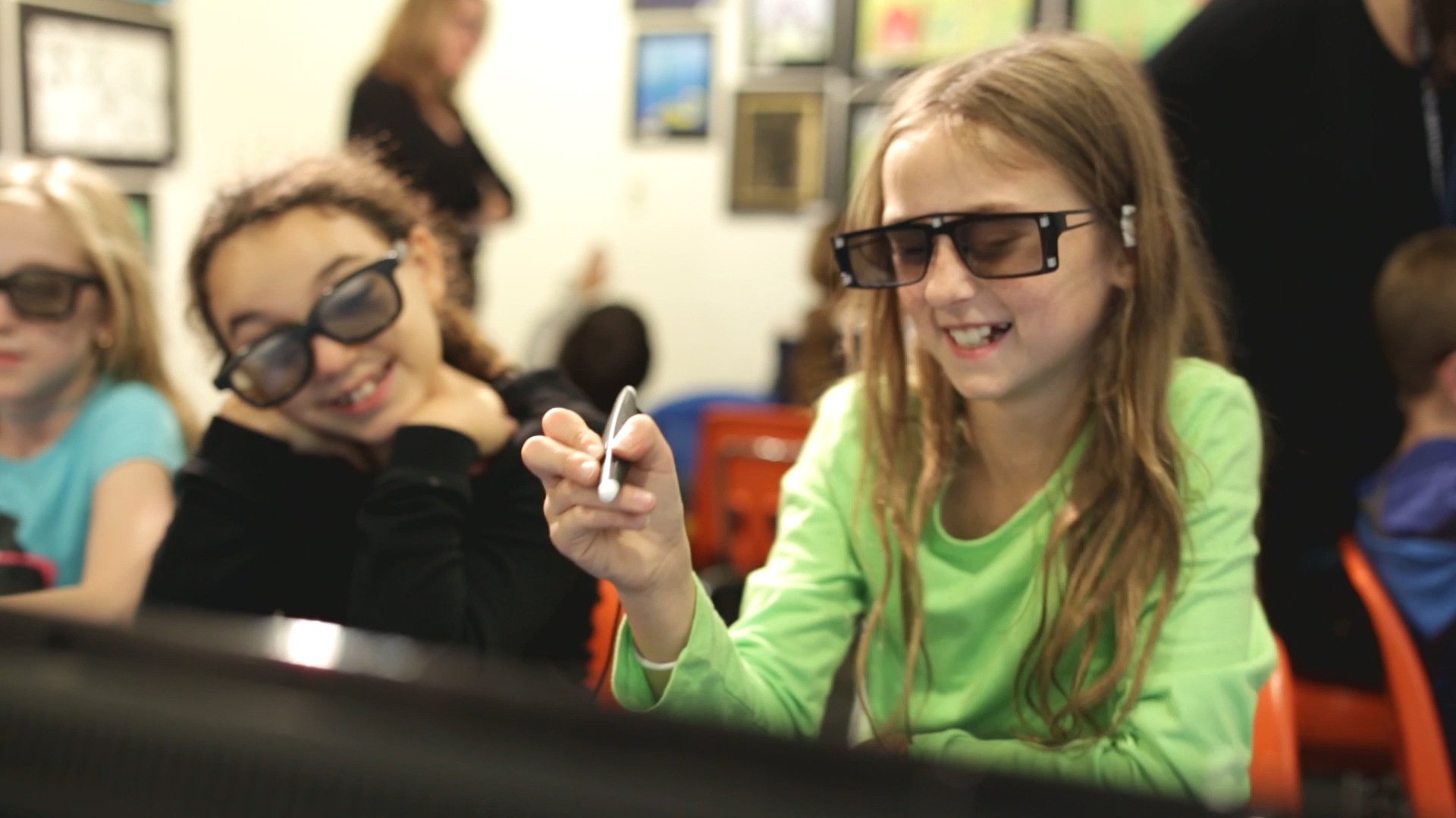 Two elementary school girls wearing 3D glasses interact with AR/VR educational content using a stylus in a classroom setting. One girl smiles while holding the stylus, engaging with immersive learning technology.