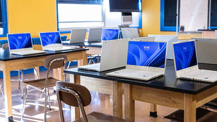 Classroom with multiple laptops displaying zSpace Inspire laptops on desks. The room has yellow walls, blue window frames, and wooden desks with brown chairs.