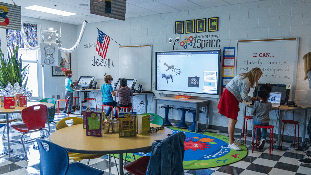 "A vibrant classroom equipped with zSpace technology, where young students engage in interactive learning. The room features a large digital screen displaying 3D animal models, individual computer stations, and colorful seating. A teacher assists a student at a computer, while others work independently. The classroom is decorated with educational posters, an American flag, and creative designs, fostering a collaborative and innovative learning environment.