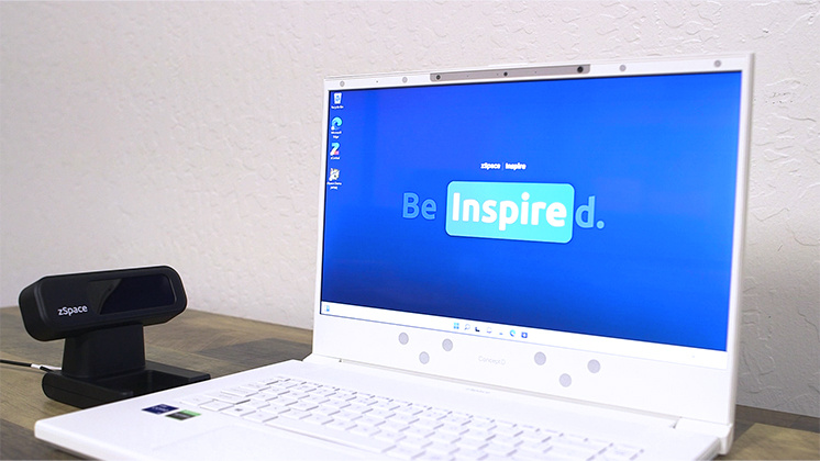 A zSpace laptop displaying the 'Be Inspired' screen with a zSpace tracking device placed beside it on a wooden desk.