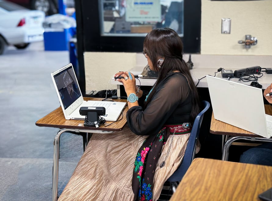 A person is sitting at a desk using a laptop with a stylus in hand. The person is wearing a traditional outfit with intricate designs. Another person is seated next to them, also using a laptop. The setting appears to be a classroom or workshop with cars visible through a window in the background. The scene suggests a blend of modern technology and traditional attire, highlighting the integration of cultural heritage with contemporary education or work environments.