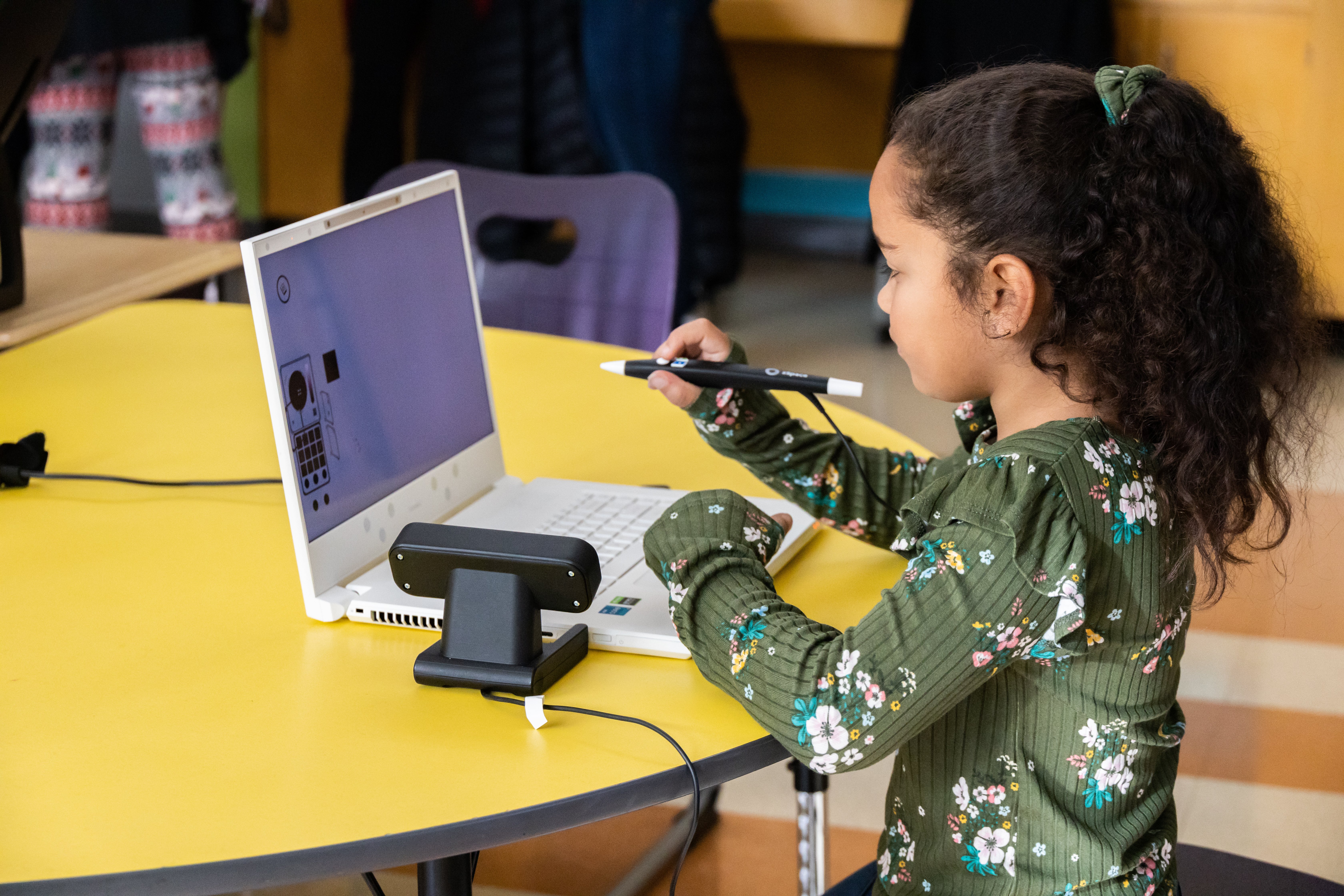 Child using a white laptop and digital pen at a yellow table, focused on educational or creative activity.