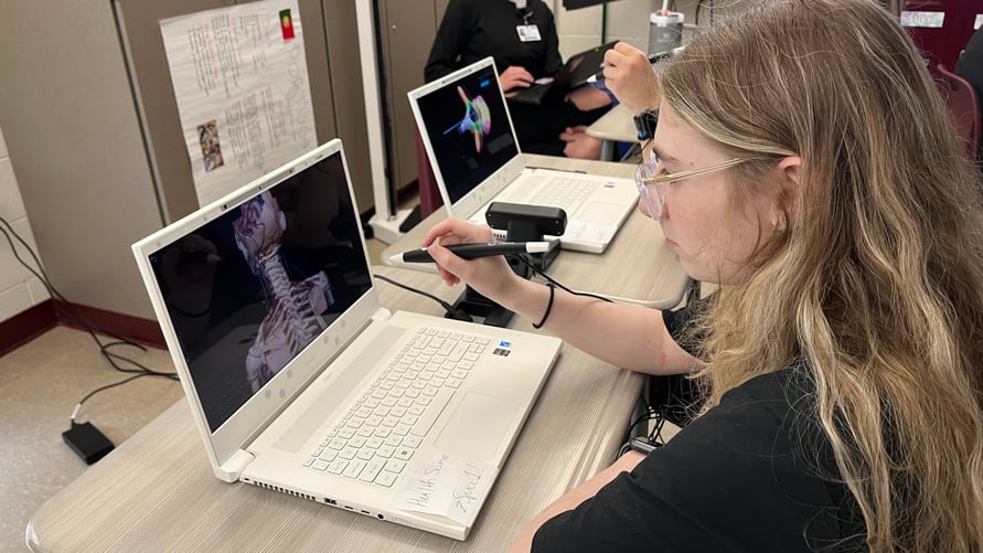 "Student using a stylus to interact with a 3D anatomical model displayed on a zSpace laptop, focusing on a detailed rendering of a human skeleton. Another student in the background engages in a similar setup, highlighting the interactive and collaborative learning environment facilitated by zSpace's augmented reality technology.