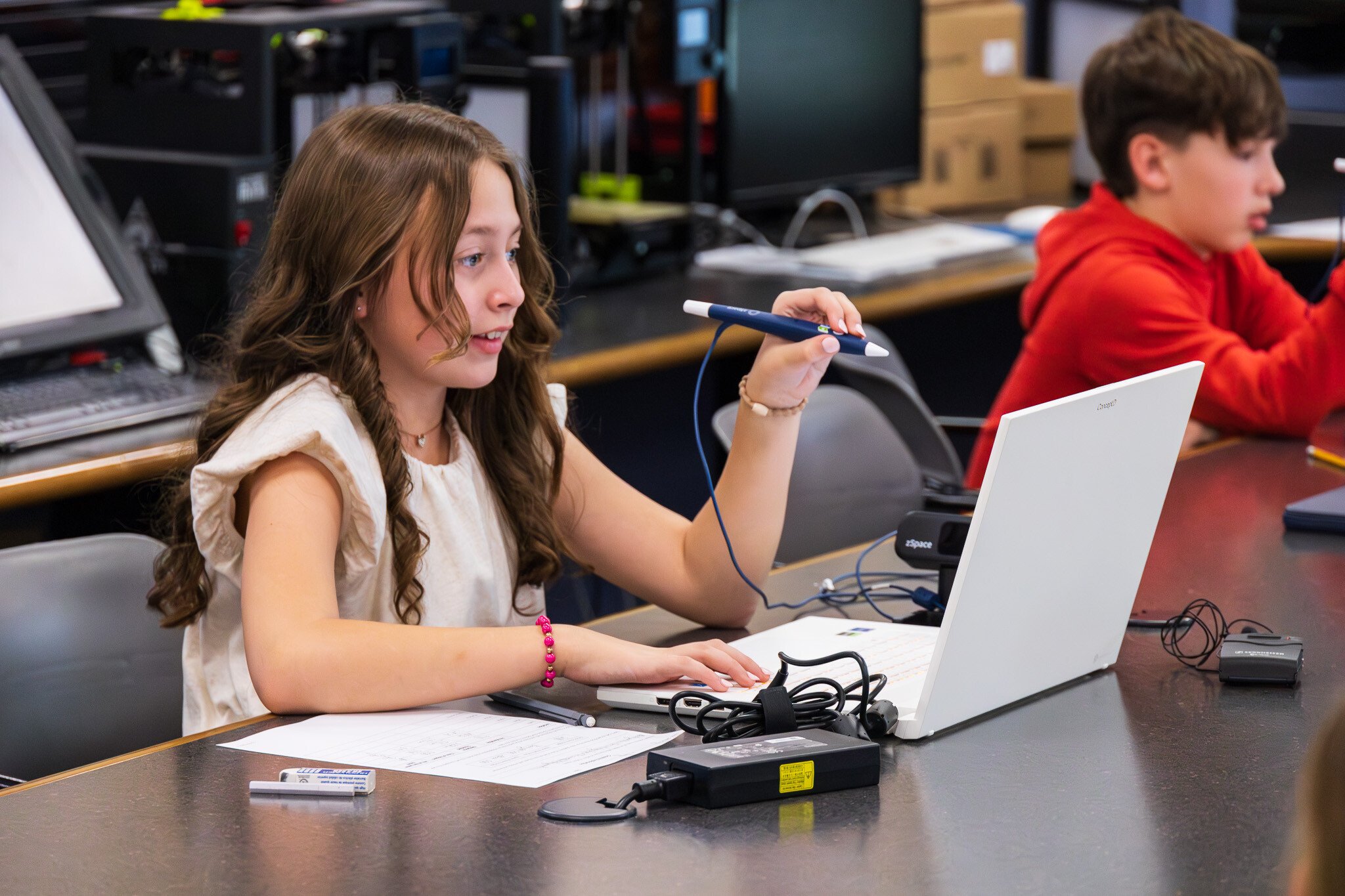 A student uses a white laptop with a stylus pen, working on a digital learning activity. In the background, another student in a red hoodie is also engaged at their desk. The classroom setting includes multiple computers and electronic devices, highlighting the integration of technology in education.