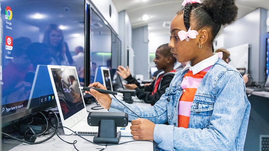 Young student uses a stylus to draw on a zSpace laptop in a classroom setting. The screen displays a simple drawing, and other students are also engaged with zSpace devices in the background. The classroom is equipped with multiple zSpace laptops and large monitors, creating a modern, technology-enhanced learning environment.