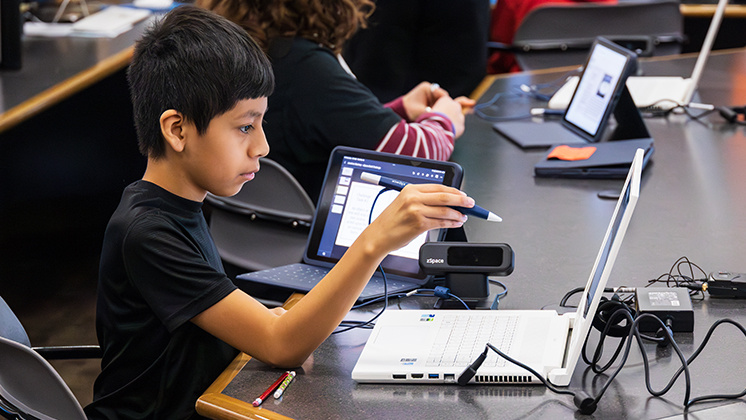 A student uses a stylus to interact with a zSpace Inspire laptop and tablet in a classroom setting. The student is engaged in a hands-on learning activity, utilizing zSpace's augmented and virtual reality technology. Other students are visible in the background, also working with similar devices.