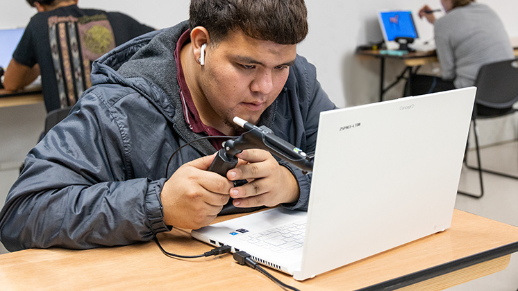 "A person wearing a gray jacket and earphones is using a zSpace laptop and a stylus for interactive learning. The background shows other individuals also working on laptops at desks, suggesting a classroom or lab environment.