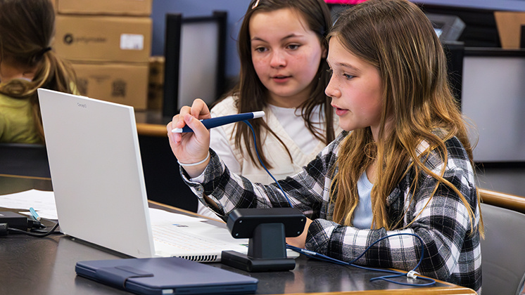 "Students using zSpace laptop and stylus for interactive learning in a classroom setting. The image shows a student holding a stylus and engaging with a zSpace laptop, highlighting the use of advanced technology in education. The classroom environment includes other students and educational materials.