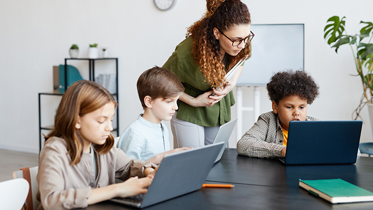 A teacher assists three students working on laptops in a modern classroom setting. The students are seated at a table with their laptops open, while the teacher leans over to provide guidance. The classroom features shelves, plants, and a whiteboard in the background.
