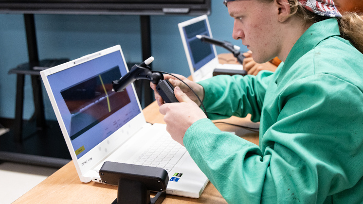 A person in a green lab coat uses a stylus to interact with a 3D simulation on a zSpace laptop. The screen displays a virtual environment, likely for educational or training purposes. Multiple laptops with styluses are present, indicating a collaborative or classroom setting.