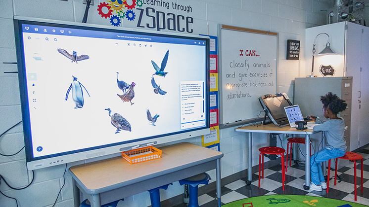 A student interacts with a zSpace virtual reality system in a classroom. The large screen displays various 3D models of birds, while the student uses a stylus and a laptop to engage with the content. The classroom features educational posters and a whiteboard with the learning objective: 'I CAN... classify animals and give examples of appropriate habitats and adaptations.' This highlights the use of advanced technology to enhance learning experiences in educational settings.