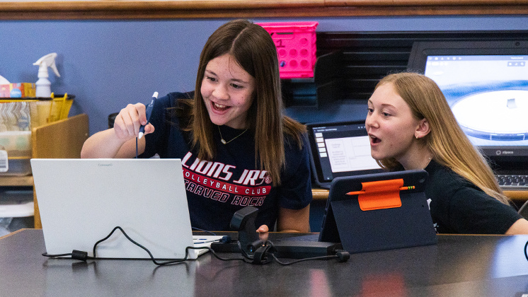 Two students using zSpace technology in a classroom setting. One student is holding a stylus and interacting with a zSpace Inspire laptop, while the other is looking at a tablet. The scene highlights the use of interactive and immersive learning tools in education, showcasing the advanced capabilities of zSpace technology in enhancing student engagement and understanding.