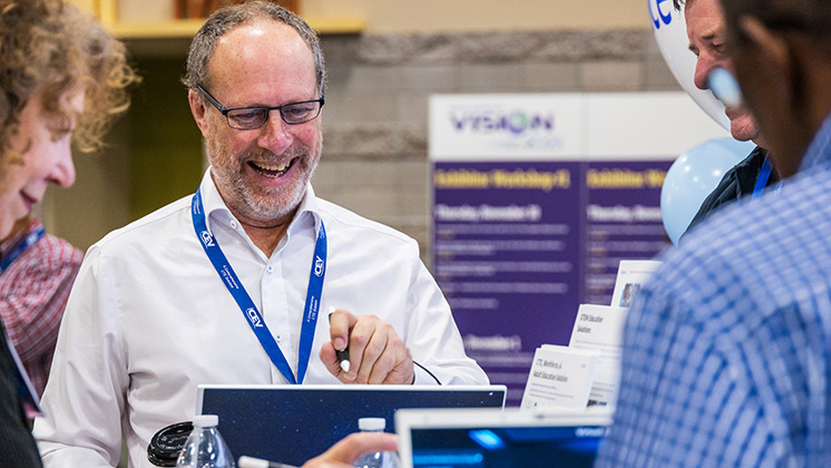 A group of people at a technology conference booth, engaging in a discussion. One person is pointing at a laptop screen, wearing a white shirt and a blue lanyard with "zSpace" branding. The background features a purple banner with the word "VISION" and other text.