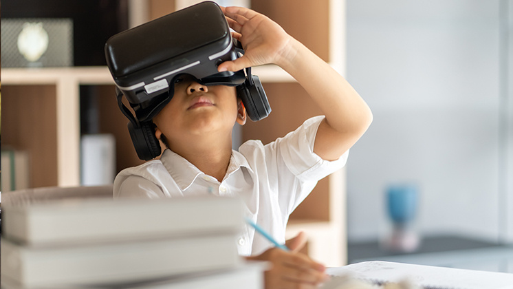 "A child wearing a virtual reality headset, sitting at a desk and holding a pencil while engaging with a notebook. In the foreground, there is a stack of books, and the background shows shelves and a blue object, slightly blurred.