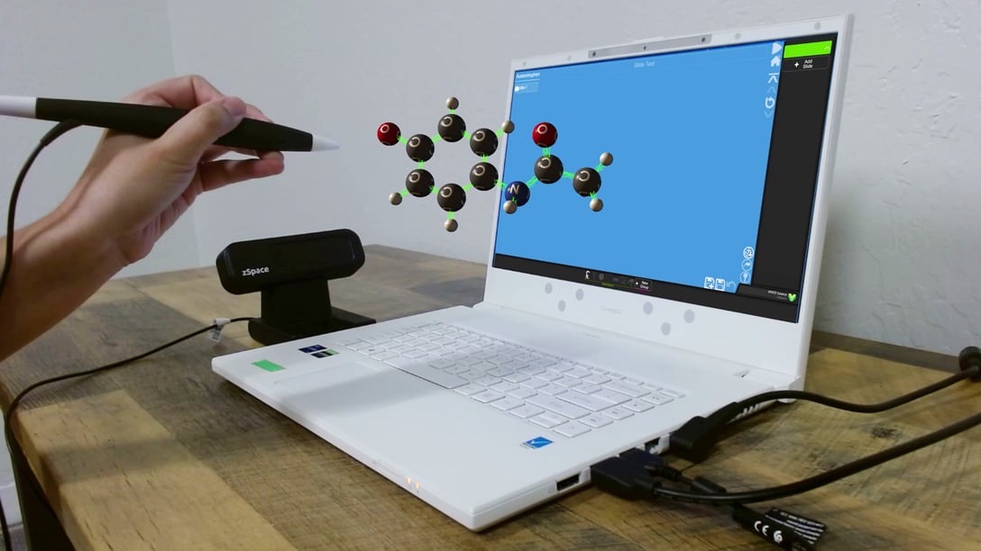 A person uses a stylus to interact with a 3D molecular model displayed on a zSpace laptop. The laptop screen shows a blue interface with the molecular structure extending into the air, demonstrating zSpace's augmented reality capabilities for educational purposes.