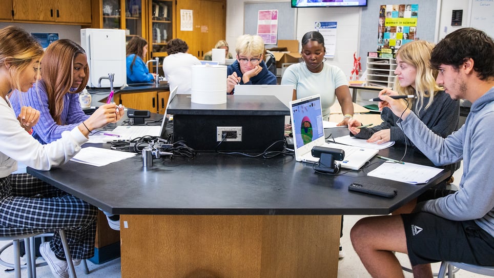 Students engaged in a collaborative science lesson using zSpace technology in a classroom. The image shows a group of students sitting around a hexagonal table, working on laptops and using styluses to interact with 3D models displayed on the screens. The classroom is equipped with scientific equipment and educational posters, emphasizing a hands-on learning environment. This setup highlights the integration of immersive technology in education, fostering interactive and engaging learning experiences.