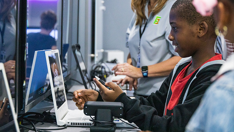 A student using a zSpace virtual reality computer in a classroom setting, guided by an instructor. The student is holding a stylus and interacting with 3D content on the screen, showcasing the immersive learning experience provided by zSpace technology.