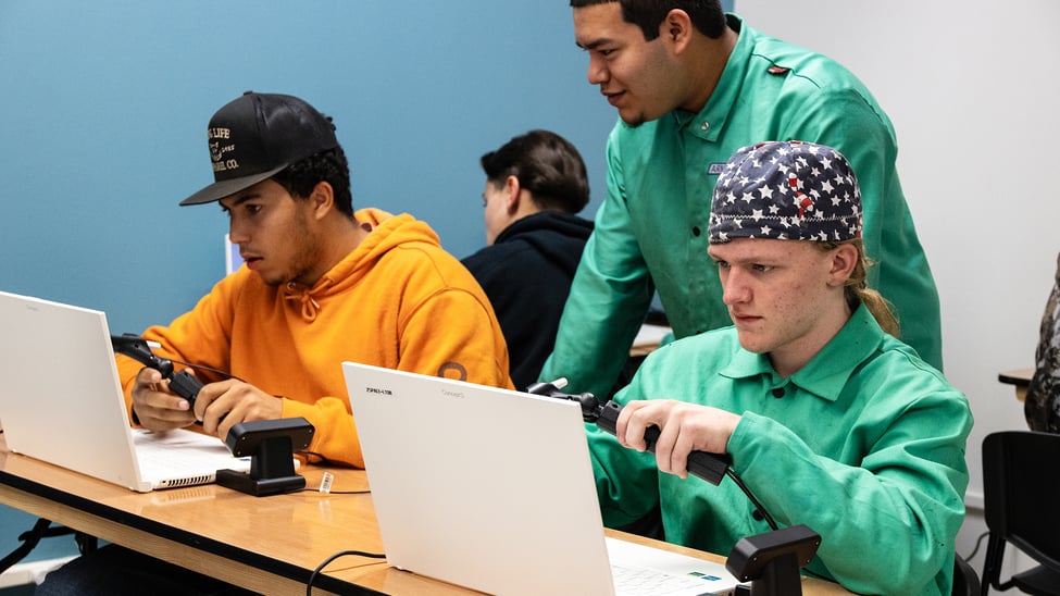 "Students using zSpace laptops and handheld controllers in a classroom setting. One student wears an orange hoodie and a black cap, while another wears a green lab coat and a star-spangled bandana.