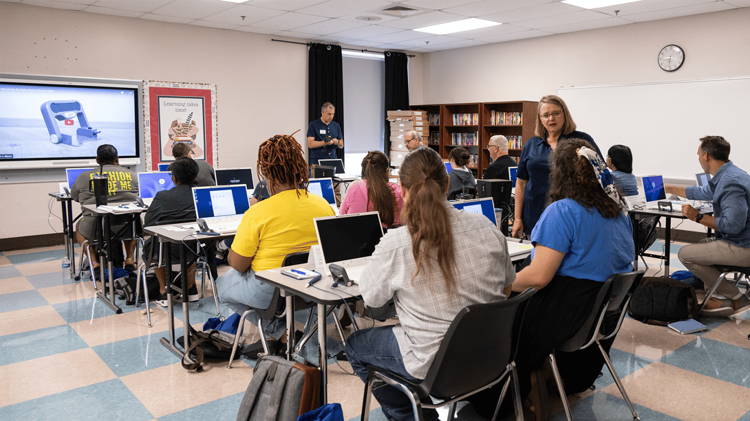 Classroom filled with adult students using zSpace laptops for an interactive learning session, guided by an instructor at the front. A 3D model is displayed on a large screen, showcasing modern technology and hands-on learning in education.