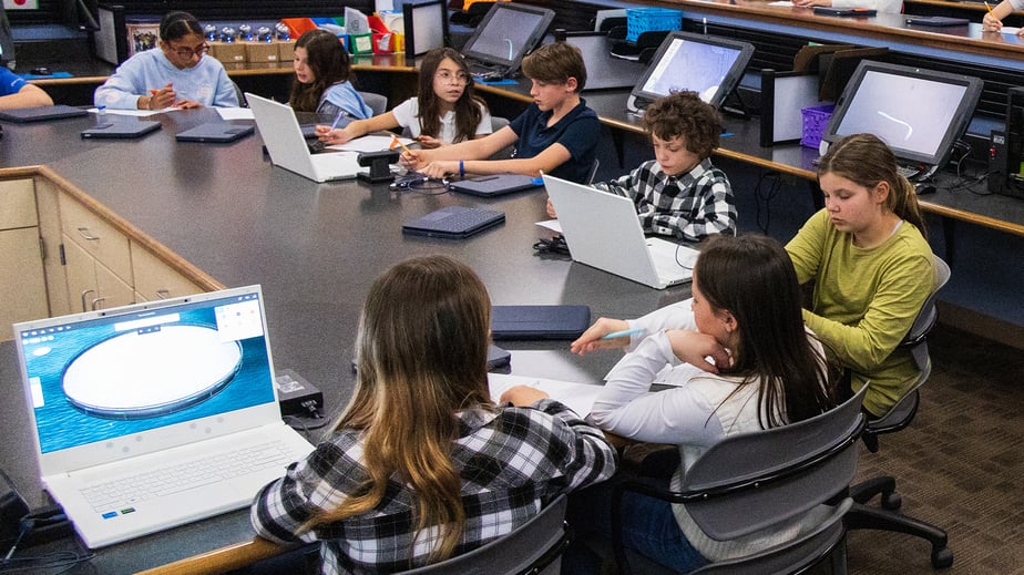 Group of students around a table engaged in collaborative learning using zSpace laptops and tablets in a classroom setting. The scene highlights hands-on educational activities with various digital devices and tools, showcasing the integration of advanced technology in modern education.