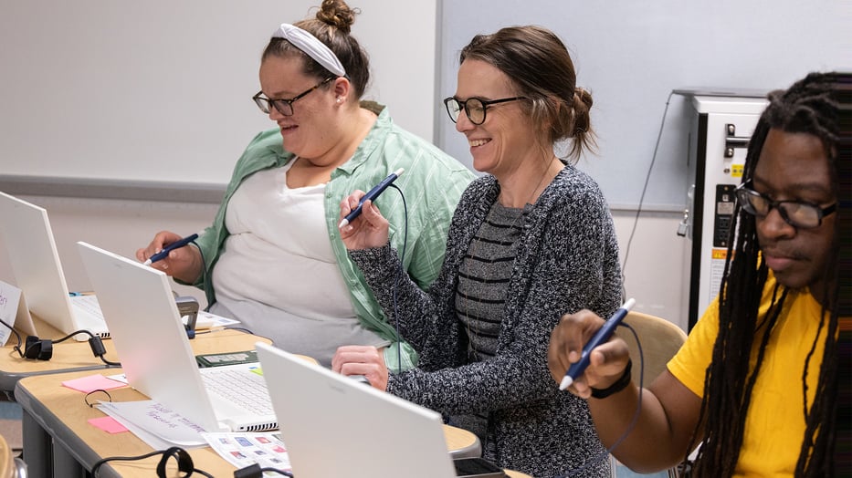 "Three individuals seated at a table using zSpace laptops and styluses in a classroom setting. They are engaged in an interactive learning activity, showcasing the use of zSpace technology in education. The table is equipped with educational materials such as notebooks and printed guides.