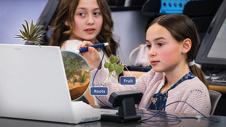 Two students using a zSpace laptop and stylus to interact with a 3D model of a plant. The model highlights various parts of the plant, including roots and fruit.