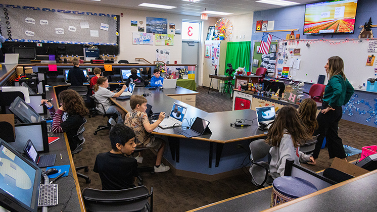 Classroom filled with students using zSpace laptops and VR headsets. The students are engaged in interactive learning activities, with a teacher assisting them. The classroom is equipped with various educational tools and decorations, including a green screen, posters, and an American flag.