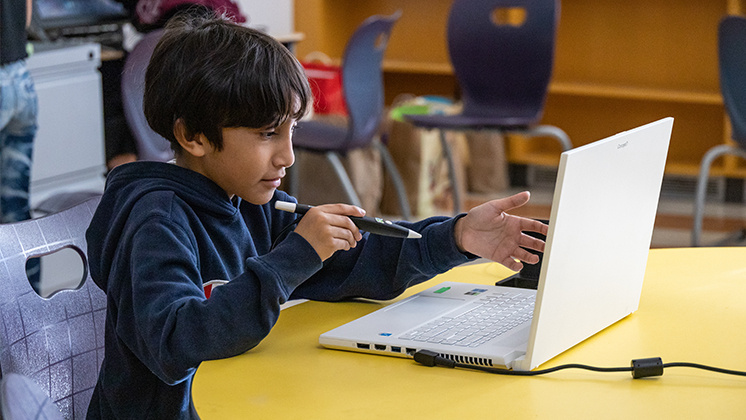 A student using a zSpace Inspire laptop at a yellow table in a classroom, holding a pen and gesturing while engaging in an online lesson or virtual meeting. The classroom features several chairs and shelves in the background, highlighting the integration of digital devices in modern education.