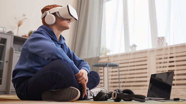 A young boy wearing a virtual reality headset and headphones sits cross-legged on the floor in a modern living room. They are dressed in a blue hoodie and jeans, with a laptop and additional headphones placed on the floor nearby.