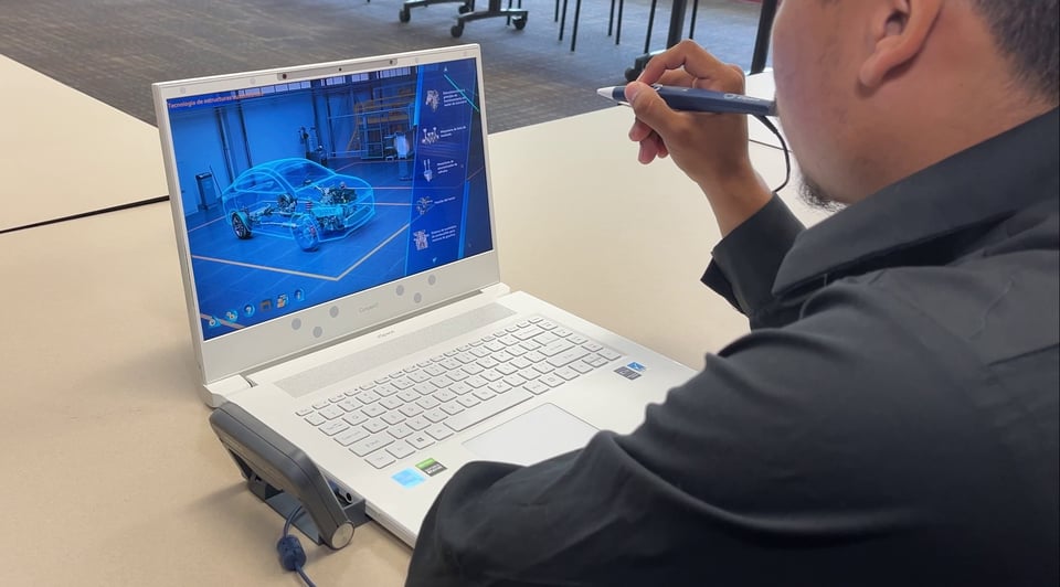 Person in a black shirt interacting with a white laptop displaying a detailed 3D model of a car's internal structure, set in a room with tables and chairs.