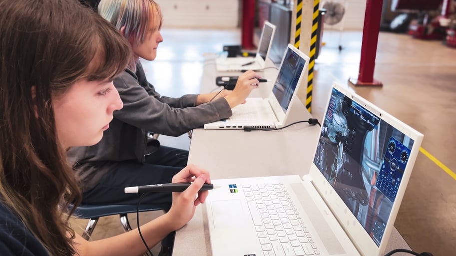 Female high school students using stylus pens to interact with 3D simulations on zSpace laptops in a classroom setting. The image shows students seated at a table, each using a stylus pen to interact with 3D simulations displayed on zSpace laptops. The classroom environment includes safety-striped pillars and various equipment in the background, indicating a hands-on learning space.