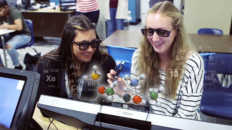 Two students using zSpace's augmented reality technology to study a 3D molecular model in a classroom. The students are wearing 3D glasses and interacting with the model using a stylus, with elements from the periodic table displayed around them.