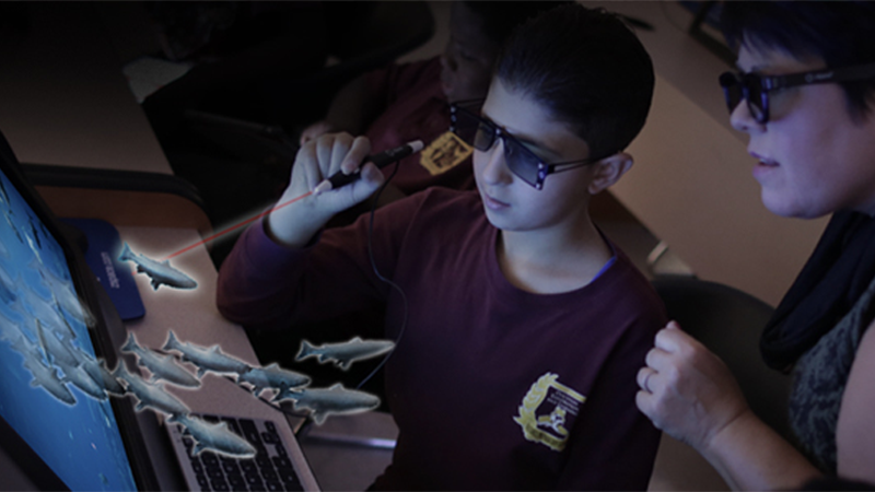 A student uses a stylus to interact with a 3D simulation of fish on a zSpace laptop, while an instructor observes.